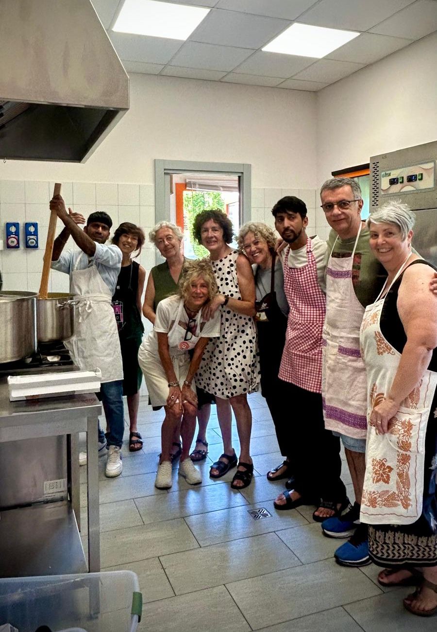 Volontarie e volontari in grembiule sorridono in una grande cucina; uno di loro alza il mestolo di legno.