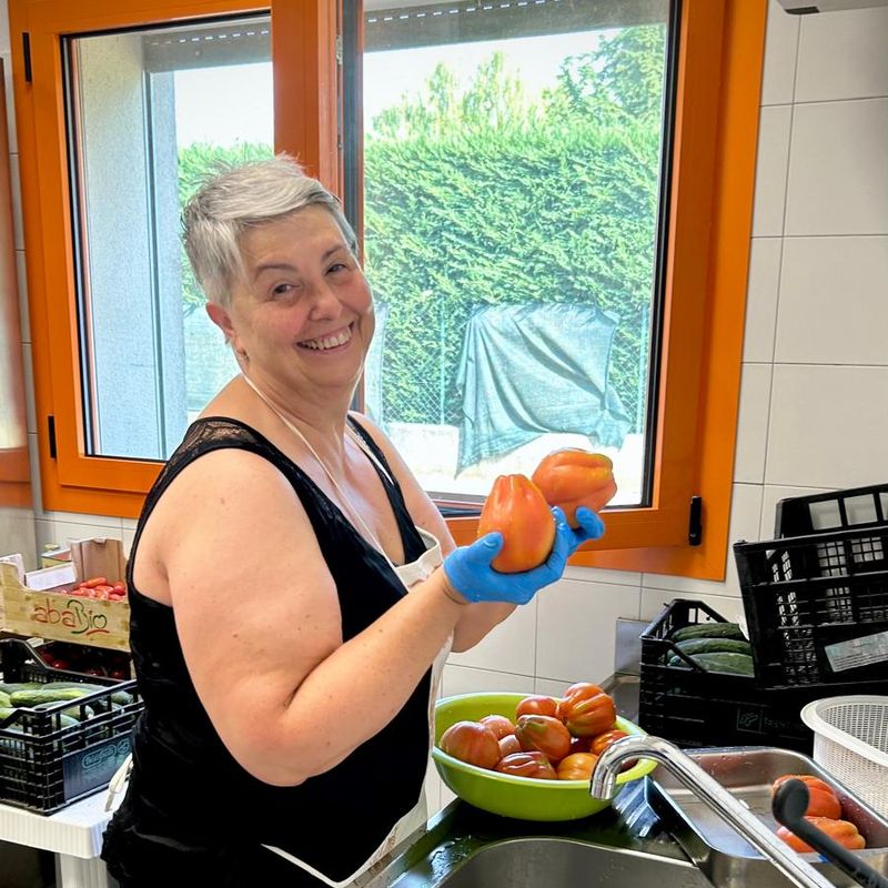 Una volontaria sorride mostrando due pomodori, accanto al lavello della cucina.