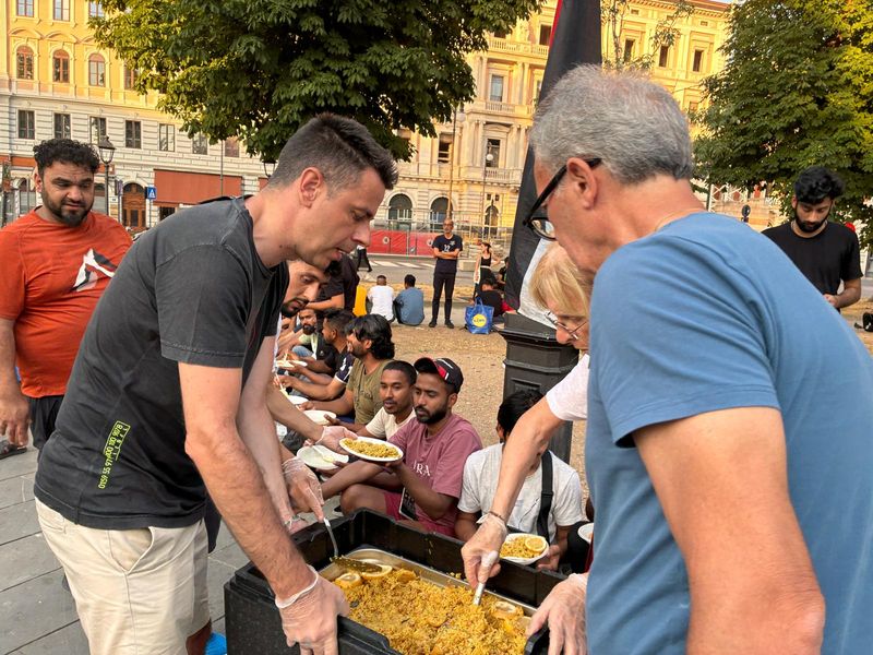 Due volontari servono riso con verdure e limone dal contenitore termico; intorno, i ragazzi aspettano col piatto in mano.