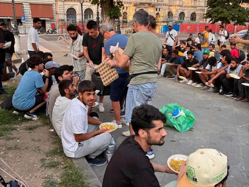 Ragazzi seduti in cerchio mangiano dai piatti; un volontario passa fra loro con le uova.